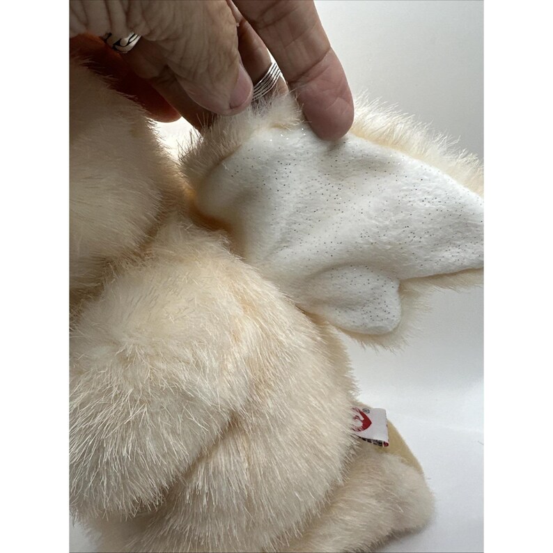 May include: A cream-coloured plush toy with white, glittery wings. The toy has a soft, furry texture and a small tag attached. The wings are made of a smooth, padded material.