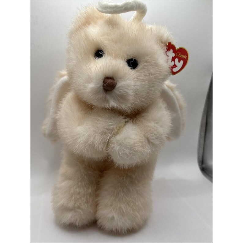 May include: A cream-coloured plush teddy bear with black eyes, a brown nose, and white wings. The bear has a halo and a red tag that says "Classic". The bear is standing with its paws together.