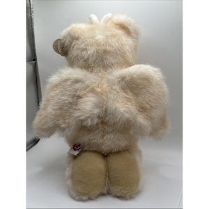 May include: A cream-coloured teddy bear with large, feathery wings. The bear has soft, textured fur and a tag attached to its ear. The bear's feet are a lighter shade of cream. A small red tag is attached to the bear's rear.