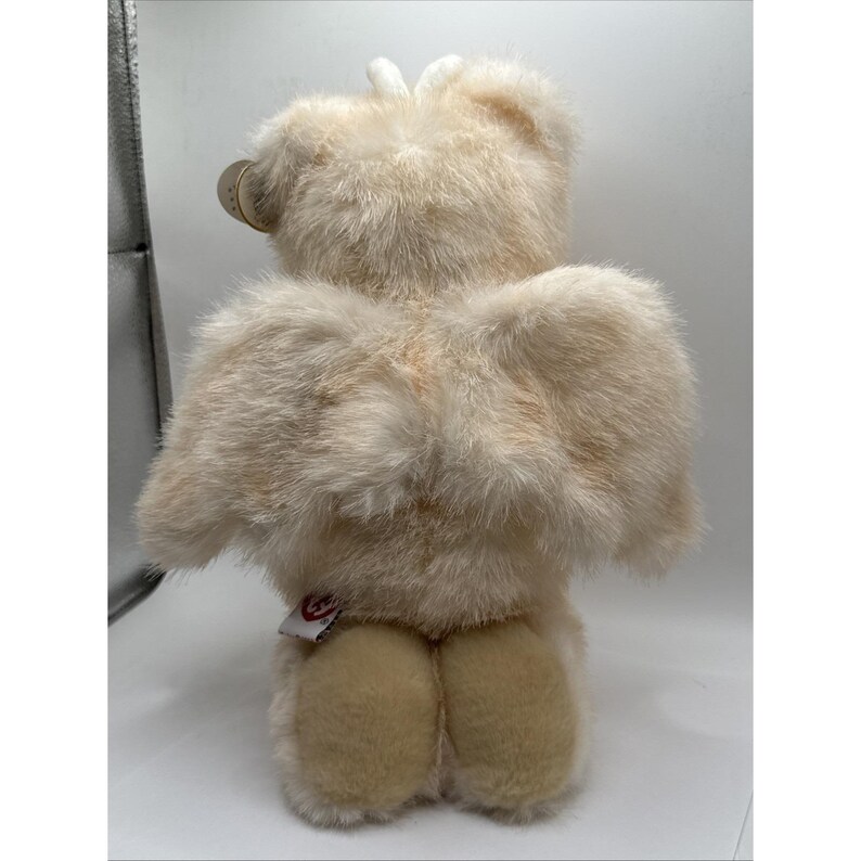 May include: A cream-coloured teddy bear with large, feathery wings. The bear has soft, textured fur and a tag attached to its ear. The bear's feet are a lighter shade of cream. A small red tag is attached to the bear's rear.