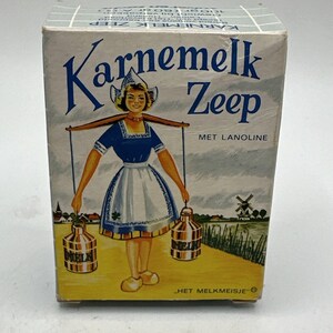 Puede incluir: Una caja vintage de jabón Karnemelk Zeep con lanolina. La caja presenta una ilustración colorida de una lechera holandesa que lleva dos bidones de leche en un yugo. El texto en la caja dice "Karnemelk Zeep met Lanoline" y "Het Melkmeisje".