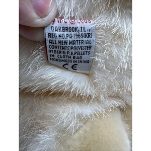 May include: Close-up of a plush toy tag. The tag is white with red text and includes the text "OAKBROOK, IL" and "ALL NEW MATERIAL". The toy is a light cream colour and has a soft, fluffy texture.