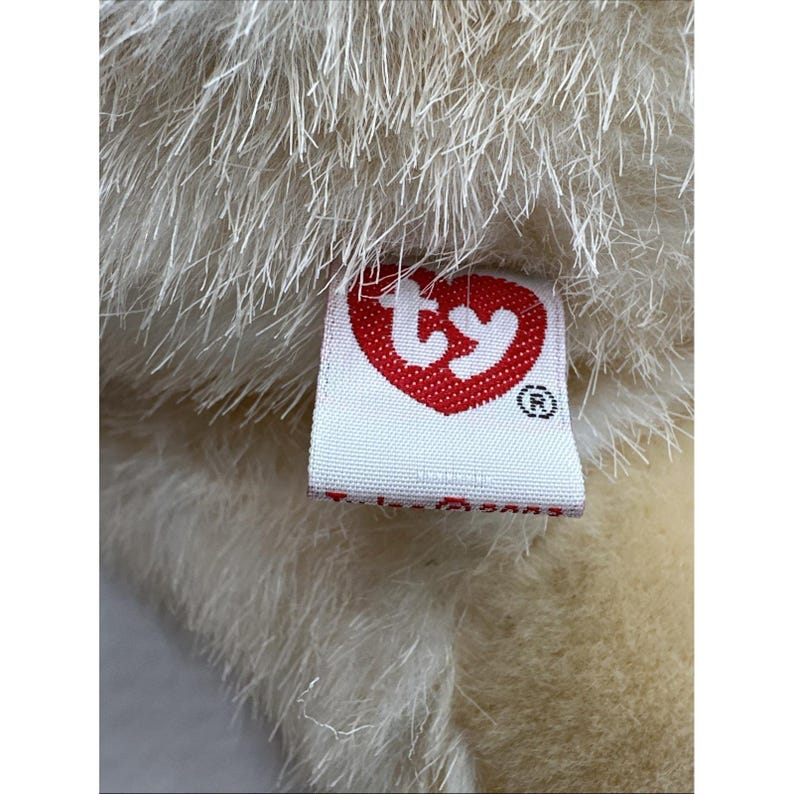 May include: Close-up of a plush toy with cream-coloured fur. A white tag with a red heart-shaped logo and the word "Ty" is attached to the toy. The tag also has a registered trademark symbol.