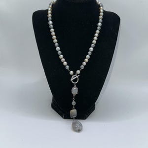 May include: A pearl necklace with a silver toggle clasp and a pendant featuring gray and white stones. The necklace is displayed on a black mannequin, showcasing its design and color variations.