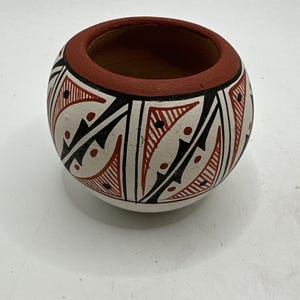 Puede incluir: Una pequeña olla redonda de cerámica con una abertura ancha. La olla está pintada con una base blanca y presenta un patrón repetitivo de diseños geométricos negros y rojos. El borde es de color marrón rojizo sólido. La olla es probablemente hecha a mano.