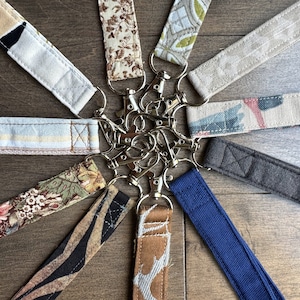 May include: A collection of fabric keychains with different patterns and colours, including floral, geometric, and striped designs. Each keychain has a silver metal key ring.