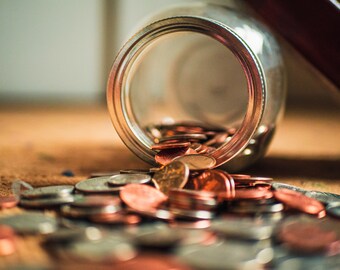 May include: A glass jar tipped over, spilling out a collection of copper and silver coins. The coins are scattered on a brown surface.