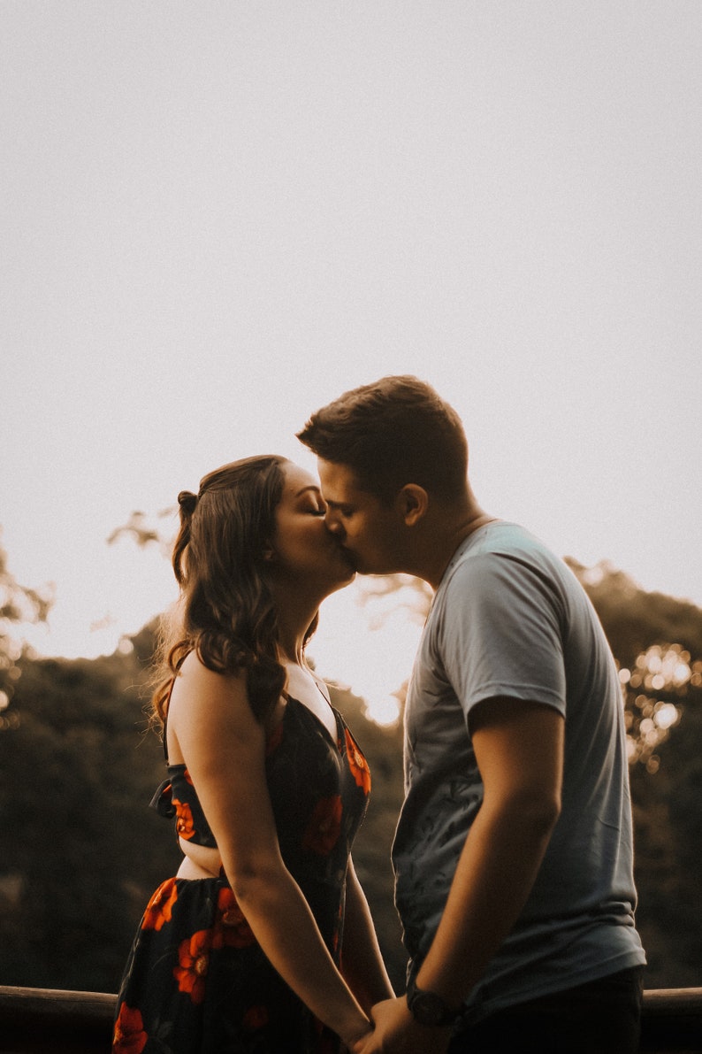 May include: A couple kissing outdoors. The woman is in a black dress with red floral patterns. The man wears a light blue t-shirt. They hold hands, with a blurred background of trees and sky.