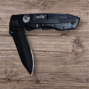 Puede incluir: Cuchillo plegable negro con una hoja parcialmente dentada. El cuchillo presenta un logotipo de águila plateada y el texto "ONEAGE" en el mango. La hoja está abierta, revelando un filo afilado. El cuchillo está sobre un fondo de madera.
