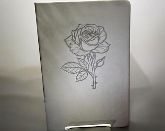 May include: A rectangular notebook with a dark gray cover featuring a centered, detailed line art illustration of a single blooming rose with leaves and a stem. The cover has a smooth, matte finish with subtle light reflections across the surface.