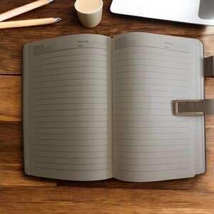 May include: An open, lined notebook with a brown leather cover and a metal clasp. The pages are cream-colored and ready for writing. Pencils and a small cup are visible on the wooden surface.