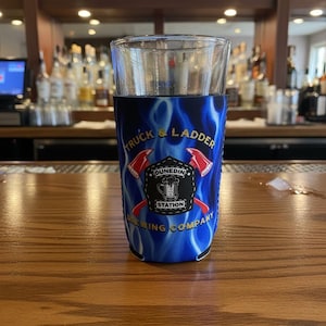 May include: A pint glass with a blue koozie. The koozie has the text "Truck & Ladder Dunedin Station Brewing Company" and a blue flame design. The logo includes crossed axes. The glass is on a wooden bar.
