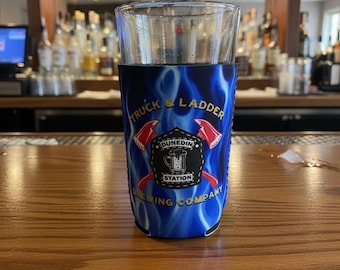 May include: A clear glass pint glass inside a blue neoprene beverage sleeve featuring a flame pattern. The sleeve displays the text Truck & Ladder Brewing Company and Dunedin Station with a graphic of two red axes and a beer mug.