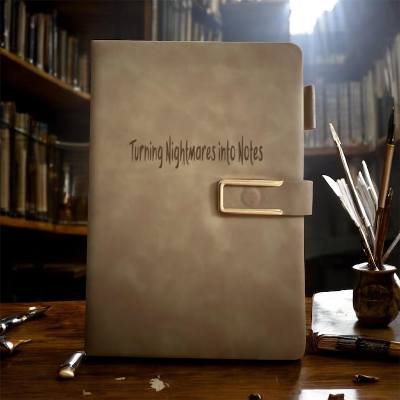 May include: A tan leather journal with the text "Turning Nightmares into Notes" embossed on the cover. The journal has a gold-colored clasp and a matching tab closure. The background features a wooden surface and a bookshelf.