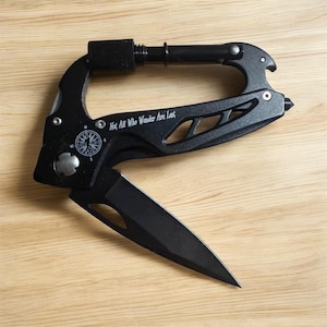Puede incluir: Mosquetón multiherramienta negro con una hoja de cuchillo plegable. El mosquetón presenta un diseño de brújula y el texto "Not All Who Wander Are Lost". Incluye un abrebotellas y una punta.