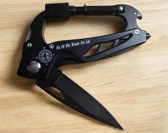 May include: A black carabiner multi-tool featuring a folding blade, a compass graphic, and the engraved text "Not All Who Wander Are Lost." The tool includes a locking gate mechanism, a pointed tip, and cut-out details along the handle frame.
