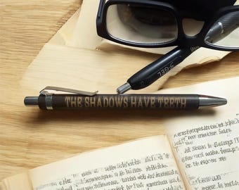 May include: A brown pen with the text "THE SHADOWS HAVE TEETH" is positioned on a wooden surface, alongside a pair of black eyeglasses and a black pen. An open book with handwritten text is in the foreground. The scene suggests a workspace or study setting.