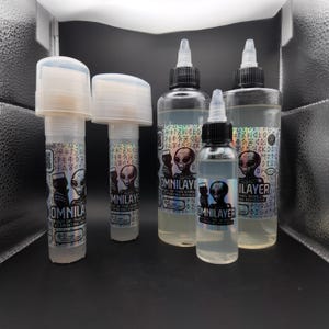 May include: A collection of clear plastic bottles and tubes, some with white caps and black nozzles. The bottles and tubes have labels with an alien graphic and the text "OMNILAYER" and "OUT OF THIS WORLD 3D PRINTING ADHESIVE".