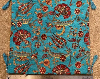 May include: A square, turquoise pillow cover with a floral pattern in red, orange, and gold. The pillow cover has tassels at each corner. A ruler is placed below the pillow cover, measuring approximately 17 inches.
