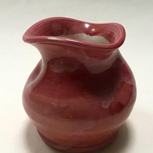May include: A small, hand-thrown ceramic jug with a wavy rim. The jug is a deep red colour with a glossy finish.