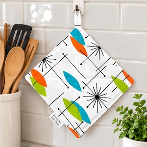 May include: A square quilted potholder featuring a white background with a repeating pattern of black starburst lines and orange, blue, and lime green leaf shapes. A small white fabric tag reads Yummy Hot Dish Tvots.