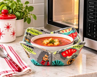 May include: A fabric bowl cozy featuring a gray background with a colorful pattern of retro-style chefs, kitchen utensils, and abstract shapes. The cozy holds a white ceramic bowl filled with red tomato soup topped with golden brown croutons.