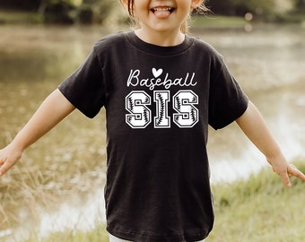 May include: A child wearing a black t-shirt with a white graphic that says "Baseball Sis" with a heart and baseball stitching. The child is standing in a grassy area near a body of water.