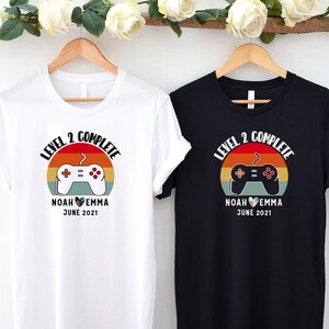 May include: Two white and black t-shirts with a retro video game controller graphic and the text "Level 2 Complete" with names and date.