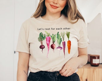 May include: A beige t-shirt with a watercolor illustration of carrots, radishes, and turnips. The text "Let's root for each other" is printed on the shirt.