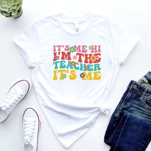 May include: White t-shirt with the text "It's me hi I'm the teacher it's me" in colorful retro font with flowers.