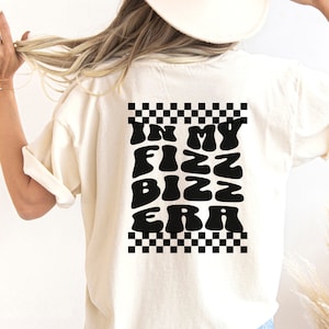 May include: A white t-shirt with a black checkered border and the text "In My Fizz Biz Era" in a retro font.