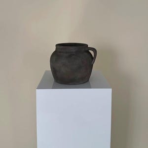 May include: A dark brown ceramic planter with a single handle. The planter is sitting on a white pedestal.