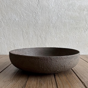 May include: A shallow, circular bowl with a dark brown, heavily textured, and matte exterior surface. The bowl features a wide, open rim and a consistent, rough, granular finish across its entire outer body, presenting a simple, functional design.