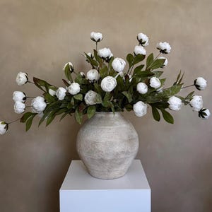 May include: A large, round, textured vase in a neutral color holds a lush arrangement of white flowers and green leaves. The vase sits on a white pedestal, creating a striking display. The flowers are in full bloom.