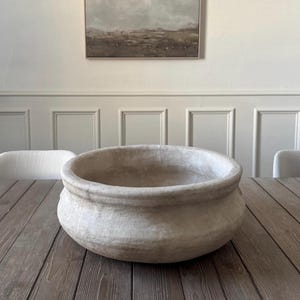 May include: A large, round, off-white bowl with a thick, rolled rim and a textured, matte finish. The vessel features a wide, deep basin and a slightly bulbous body, showcasing a weathered, earthy appearance with subtle variations in its light tone.