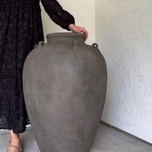 May include: Large, gray, textured ceramic vase with two small handles on each side. The vase has a wide body and a narrow neck. The vase is a neutral color and would be a great addition to any home decor.