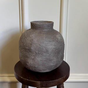 May include: A textured, gray ceramic vase with a rounded body and a short neck. The vase sits on a small, dark wooden stool with three legs. The background is a white wall with molding.