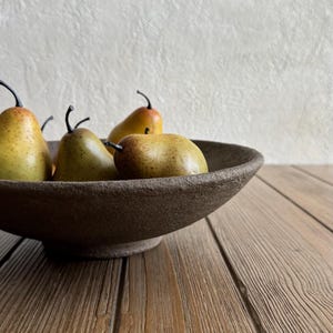 May include: A dark brown, textured, shallow ceramic bowl with a wide rim and a pedestal base. Inside the bowl are four artificial pears, each featuring a yellow-green skin with brown speckles and a thin, dark brown stem protruding from the top.