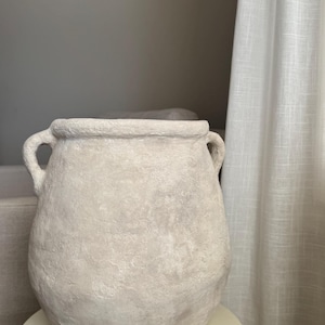 May include: A large, off-white, textured ceramic vase with two loop handles. The vase has a rounded body and sits on a small, cream-coloured pedestal. The background includes a neutral-toned wall and a white curtain.