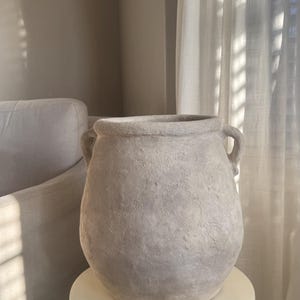 May include: A large, textured, light grey ceramic vase with two small handles. The vase sits on a white, round pedestal. The background includes a white curtain and a glimpse of a neutral-toned wall.