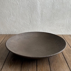 May include: A shallow, circular bowl with a dark brown, textured surface. The bowl features a wide, slightly curved rim and a rough, matte finish across its entire interior and exterior, presenting a consistent earthy tone and a simple, open form.