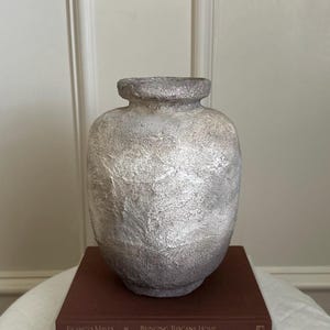 May include: A white ceramic vase with a textured surface sits on top of two books. The top book is brown and says "Bringing Tuscany Home" and the bottom book is white and says "The Art of Home".