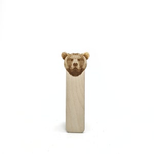 Può includere: Un segnalibro in legno con un design a testa di orso geometrico. La testa dell'orso è intagliata in modo intricato con linee dettagliate e il segnalibro è realizzato in legno di colore chiaro. Il segnalibro è rettangolare con bordi arrotondati.