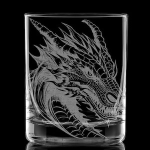 以下が含まれることがあります： ドラゴンヘッドの彫刻が施された透明なガラスのタンブラー。ドラゴンは右を向いており、鱗模様が細かく描かれています。