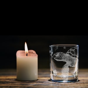 May include: A clear glass shot glass with an etched design of a sea turtle swimming with bubbles and a starfish. A lit white candle is in the background.