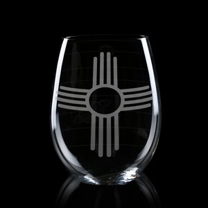 May include: A clear stemless wine glass with a New Mexico Zia symbol etched on the front. The Zia symbol is a circle with four groups of four rays extending outward. The glass has a rounded shape and is set against a black background.