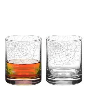 以下が含まれることがあります： 側面にクモの巣のデザインがエッチングされた透明なガラスのタンブラー2つ。1つのタンブラーは琥珀色の液体で満たされています。