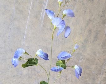 May include: A single stem of artificial blue flowers with green leaves. The delicate petals are a soft blue hue, and the leaves are a deep green. The stem is a light green color. The flower is set against a neutral background.