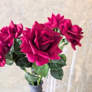 Op de afbeelding: Een boeket van diep magenta rozen met groene bladeren in een grijze vaas. De rozen staan in volle bloei en tonen hun gelaagde bloemblaadjes. De vaas heeft een eenvoudige, matte afwerking en biedt een neutrale basis voor de kleurrijke bloemen.