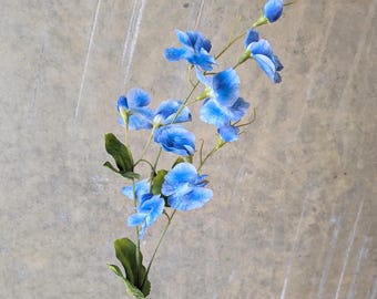 May include: A single stem of artificial blue flowers with green leaves. The flowers are a vibrant shade of blue, with darker blue accents. The stem is a light green color, and the leaves are a deep green.